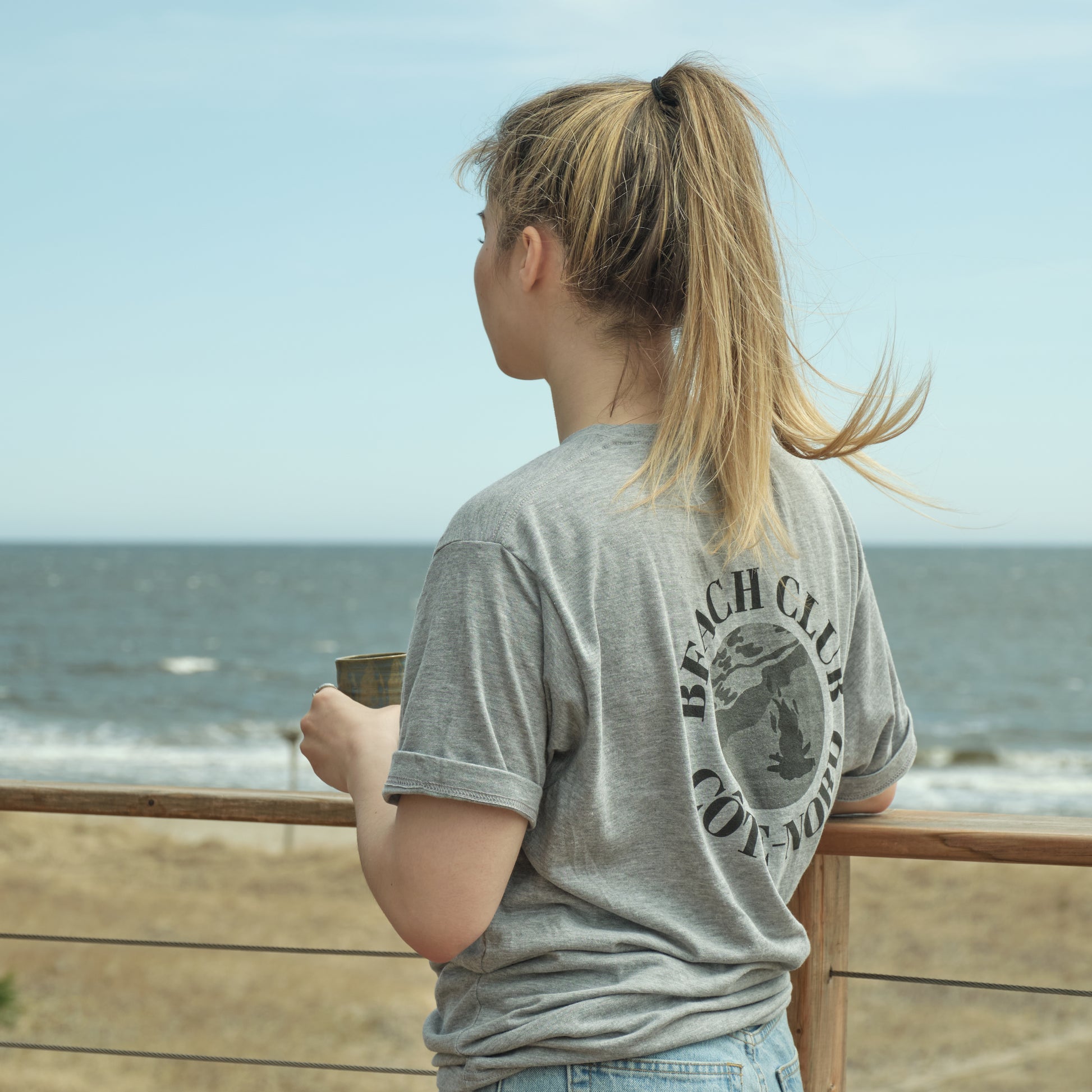 Femme prenant un café sur une galerie près de la plage portant le t-shirt beach club Côte-Nord